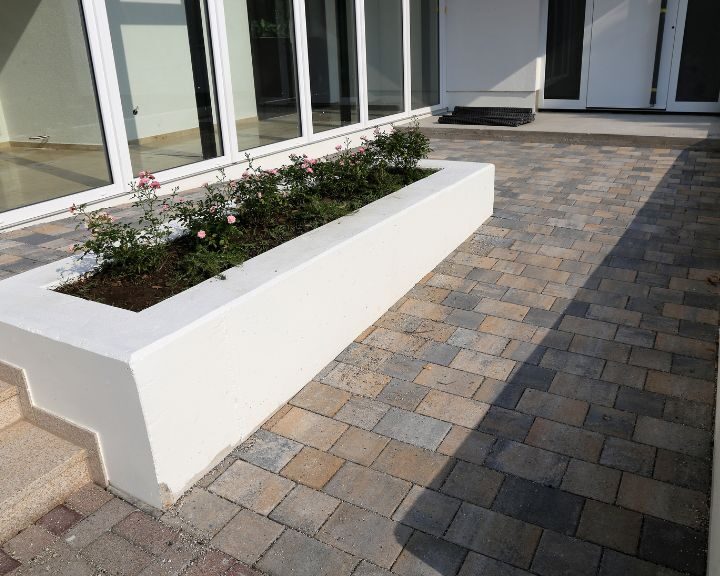 A white planter with flowers on it next to a stamped concrete house in the city.